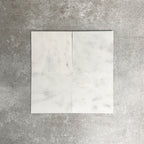 Four Fayrouz White 15.2x15.2cm natural marble tiles with a honed finish arranged together, showcasing soft white tones, subtle markings and natural variation against a neutral textured background.