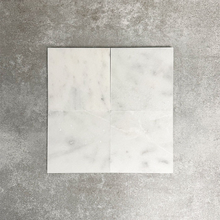 Four Fayrouz White 15.2x15.2cm natural marble tiles with a honed finish arranged together, showcasing soft white tones, subtle markings and natural variation against a neutral textured background.
