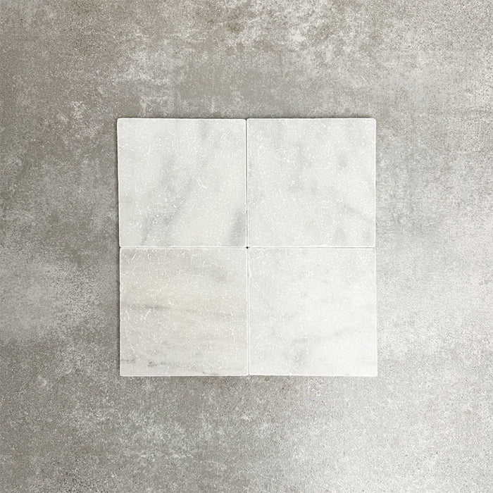 Four Fayrouz White 15.2x15.2cm natural marble tiles with a tumbled finish arranged together, showcasing soft white tones, subtle veining and natural variation against a neutral textured background.