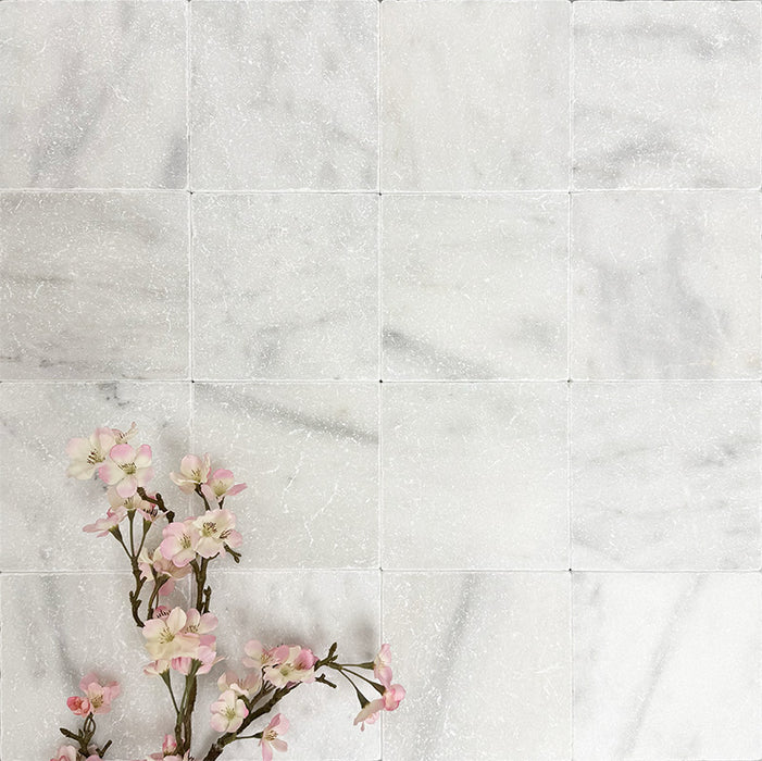 Flat lay of Fayrouz White 15.2x15.2cm natural marble tiles with a tumbled finish, featuring soft white tones and subtle natural veining, styled with delicate pink blossom branches for a calm, elegant look.