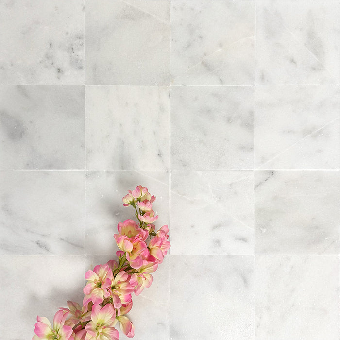Flat lay of Fayrouz White 15.2x15.2cm natural marble tiles with a honed finish, featuring soft white tones and subtle natural markings, styled with delicate pink blossom branches for a calm, elegant look.