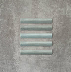 Flat lay of Zellige Aqua 3×20cm Moroccan-style ceramic bullnose tiles, showcasing the rounded edge profile, glossy blue-green glaze and subtle tonal variation on a neutral background.