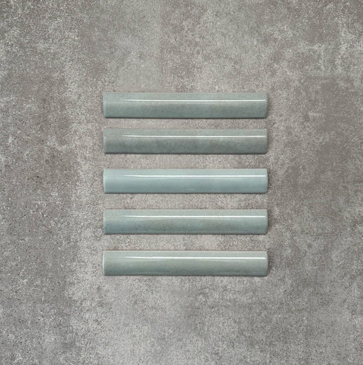 Flat lay of Zellige Aqua 3×20cm Moroccan-style ceramic bullnose tiles, showcasing the rounded edge profile, glossy blue-green glaze and subtle tonal variation on a neutral background.