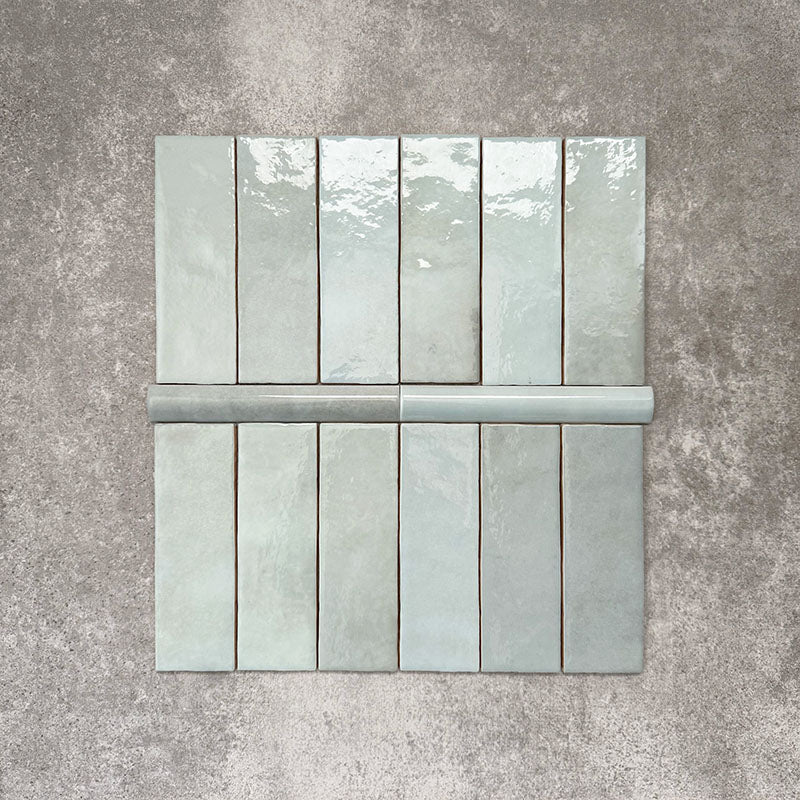 Flat lay of Zellige Aqua Moroccan-style ceramic tiles, showing 6.5×20cm brick tiles paired with a 3×20cm bullnose trim, highlighting the rounded edge detail, glossy blue-green glaze and natural tonal variation on a neutral background.