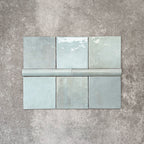 Flat lay of Zellige Aqua Moroccan-style ceramic tiles, showing 13.2×13.2cm square tiles paired with a 3×20cm bullnose trim, highlighting the rounded edge detail, glossy blue-green glaze and natural tonal variation on a neutral background.