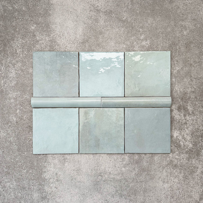 Flat lay of Zellige Aqua Moroccan-style ceramic tiles, showing 13.2×13.2cm square tiles paired with a 3×20cm bullnose trim, highlighting the rounded edge detail, glossy blue-green glaze and natural tonal variation on a neutral background.