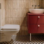 Bathroom featuring Zellige Arena Marfil Brick 6.5 × 20cm Moroccan-style ceramic wall tiles in warm beige tones, installed in a vertical brick layout behind a wall-hung toilet and red vanity unit, paired with patterned black and cream floor tiles and chrome fittings for a contemporary, characterful finish.