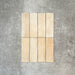Flat lay of Zellige Arena Marfil Brick 6.5 × 20cm Moroccan-style ceramic tiles, shown in a warm beige and cream colour palette with subtle tonal variation and gently uneven edges, arranged in a vertical brick layout on a textured grey background to highlight the handcrafted, zellige-effect finish.