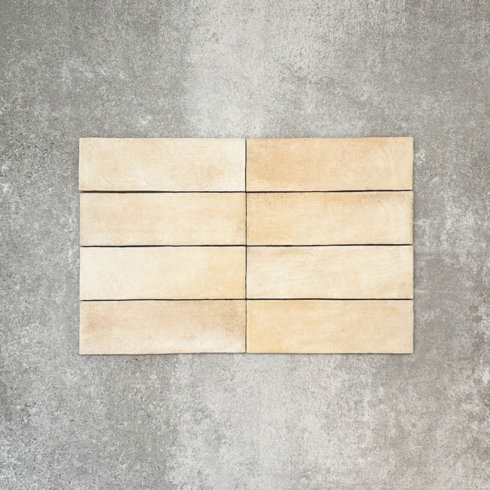 Flat lay of Zellige Arena Marfil Brick 6.5 × 20cm Moroccan-style ceramic tiles, shown in a warm beige and cream colour palette with subtle tonal variation and gently uneven edges, arranged in a vertical brick layout on a textured grey background to highlight the handcrafted, zellige-effect finish.