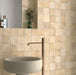 Bathroom wall featuring Zellige Arena Marfil Square 10 × 10cm Moroccan-style ceramic tiles in soft beige and sand tones, installed in a classic grid layout around a freestanding basin and brass tap, with subtle tonal variation and gently uneven edges creating an authentic handcrafted zellige-effect finish.