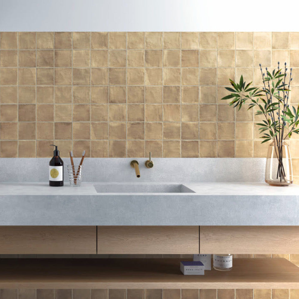 Bathroom splashback featuring Zellige Arena Marfil Square 10 × 10cm Moroccan-style ceramic wall tiles in warm beige tones, laid in a neat grid above a concrete-effect vanity with brass wall-mounted taps, with subtle colour variation and gently uneven edges adding an authentic handcrafted zellige-effect look.
