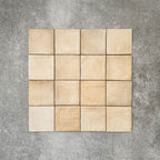 Flat lay of Zellige Arena Marfil Square 10 × 10cm Moroccan-style ceramic tiles in warm beige and sand hues, displaying subtle shade variation and softly uneven edges, arranged in a simple grid on a textured grey background to showcase the handcrafted zellige-effect surface.