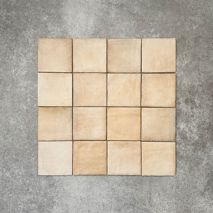 Flat lay of Zellige Arena Marfil Square 10 × 10cm Moroccan-style ceramic tiles in warm beige and sand hues, displaying subtle shade variation and softly uneven edges, arranged in a simple grid on a textured grey background to showcase the handcrafted zellige-effect surface.