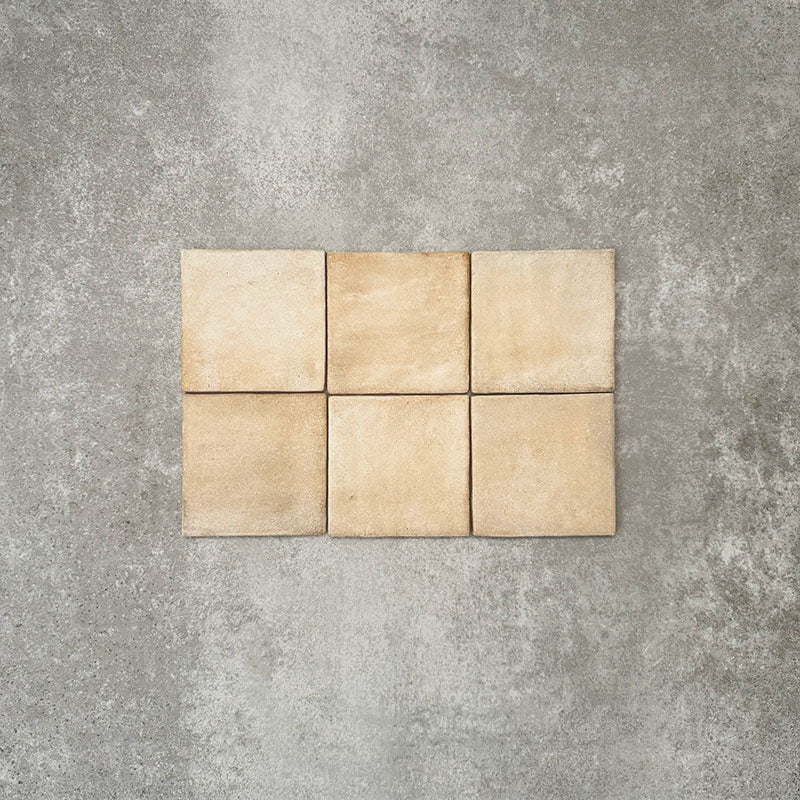Flat lay of Zellige Arena Marfil Square 10 × 10cm Moroccan-style ceramic tiles in warm beige and sand hues, displaying subtle shade variation and softly uneven edges, arranged in a simple grid on a textured grey background to showcase the handcrafted zellige-effect surface.