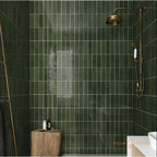 Bathroom shower area featuring Zellige Botanical Green Brick 6.5 × 20cm Moroccan-style ceramic wall tiles in deep botanical green tones, laid in a vertical brick layout, paired with vintage-style brass shower fittings and natural wood accents for a rich, atmospheric zellige-effect finish.