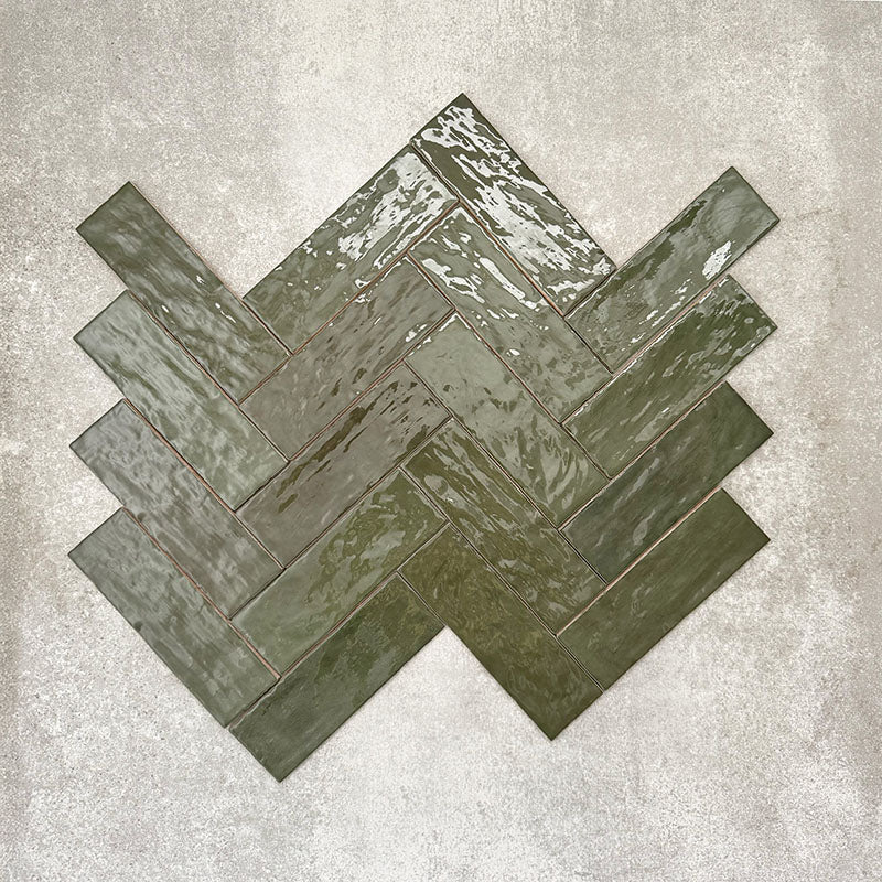 Flat lay of Zellige Botanical Green Brick 6.5 × 20cm Moroccan-style ceramic tiles in deep botanical green tones, arranged in a herringbone layout on a textured grey background, showcasing the glossy glaze, natural tonal variation and handcrafted zellige-effect finish.