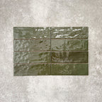 Flat lay of Zellige Botanical Green Brick 6.5 × 20cm Moroccan-style ceramic tiles in deep botanical green tones, arranged in a horizontal brick layout on a textured grey background, showcasing the glossy glaze, natural tonal variation and handcrafted zellige-effect finish.
