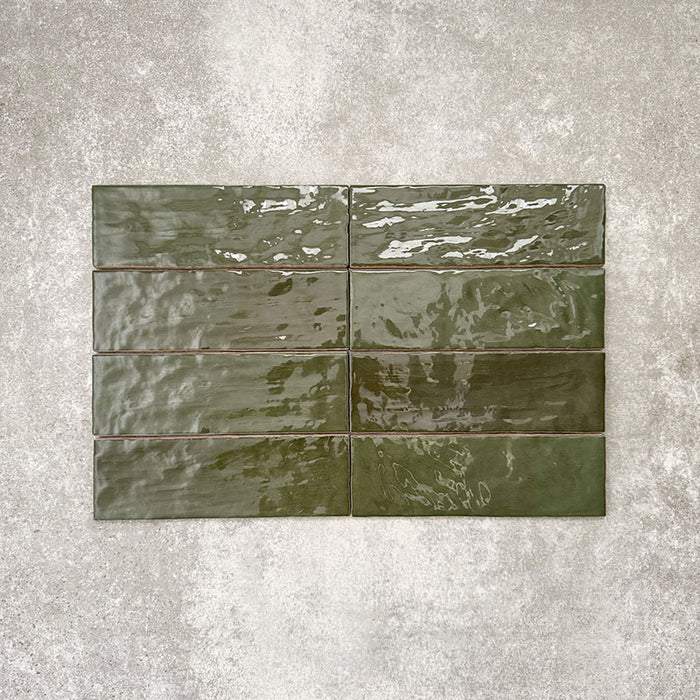 Flat lay of Zellige Botanical Green Brick 6.5 × 20cm Moroccan-style ceramic tiles in deep botanical green tones, arranged in a horizontal brick layout on a textured grey background, showcasing the glossy glaze, natural tonal variation and handcrafted zellige-effect finish.