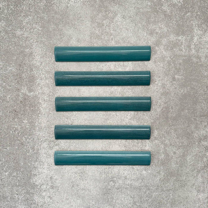 Flat lay of Zellige Peacock Green 3×20cm Moroccan-style ceramic bullnose tiles, showcasing the rounded edge profile, deep blue-green glaze and subtle tonal variation on a neutral background.