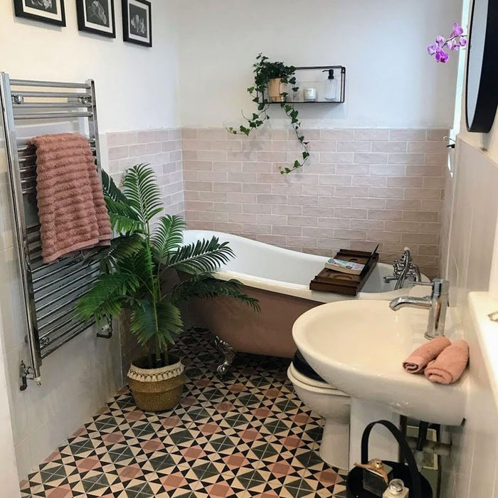 Bathroom featuring Zellige Pink Mist Brick 6.5 × 20cm Moroccan-style ceramic wall tiles in soft pale pink tones, installed in a horizontal brick layout around a freestanding bath, paired with patterned geometric floor tiles, white sanitaryware and botanical accents for a calm, characterful zellige-effect finish.