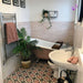 Bathroom featuring Zellige Pink Mist Brick 6.5 × 20cm Moroccan-style ceramic wall tiles in soft pale pink tones, installed in a horizontal brick layout around a freestanding bath, paired with patterned geometric floor tiles, white sanitaryware and botanical accents for a calm, characterful zellige-effect finish.