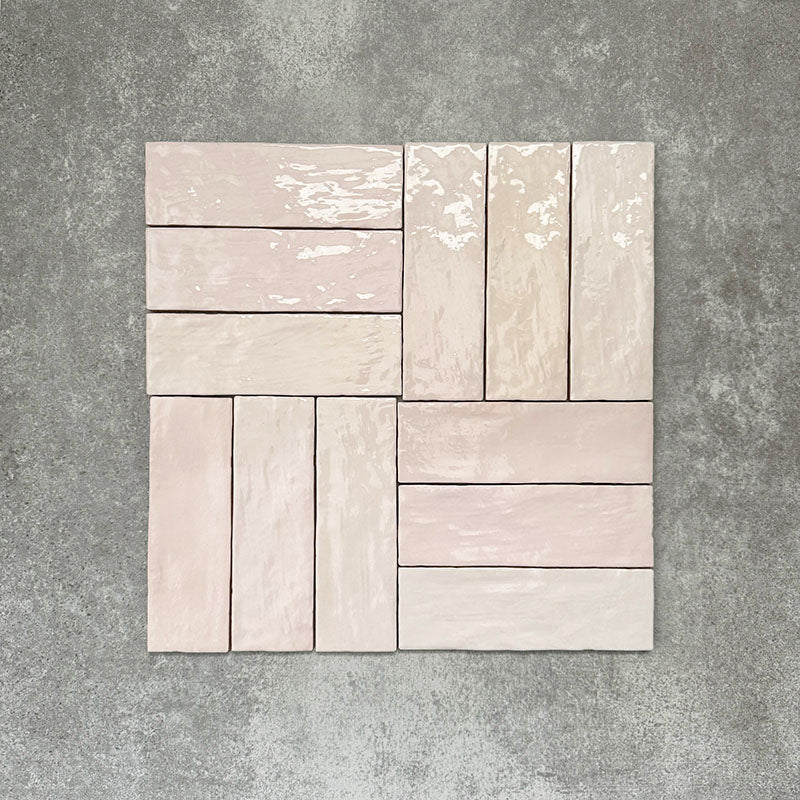 Flat lay of Zellige Pink Mist Brick 6.5 × 20cm Moroccan-style ceramic tiles in soft pale pink and blush tones, arranged in a basketweave layout on a textured grey background, showcasing the glossy glaze, gentle tonal variation and handcrafted zellige-effect finish.