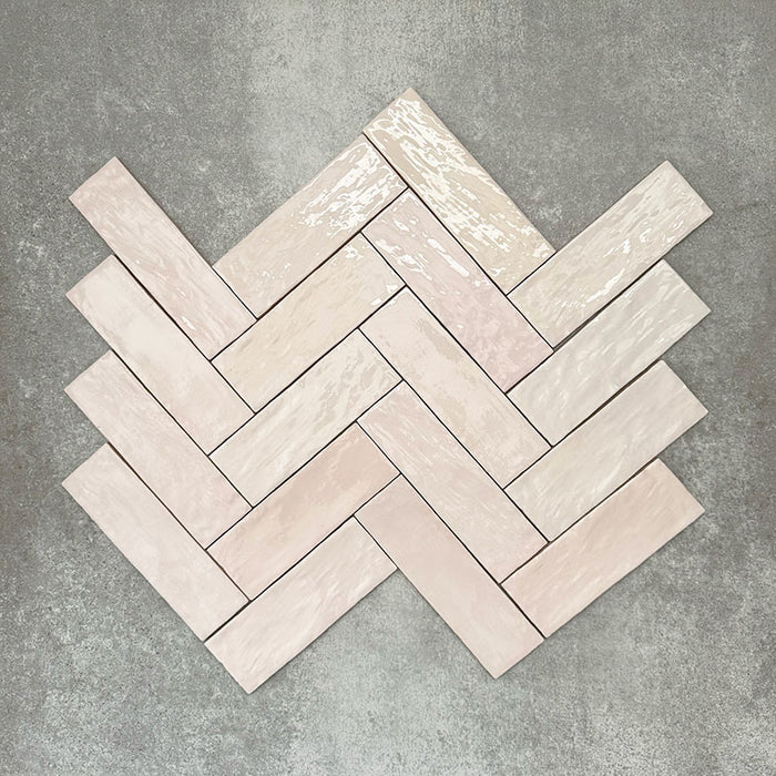 Flat lay of Zellige Pink Mist Brick 6.5 × 20cm Moroccan-style ceramic tiles in soft pale pink and blush tones, arranged in a herringbone layout on a textured grey background, showcasing the glossy glaze, gentle tonal variation and handcrafted zellige-effect finish.