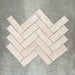 Flat lay of Zellige Pink Mist Brick 6.5 × 20cm Moroccan-style ceramic tiles in soft pale pink and blush tones, arranged in a herringbone layout on a textured grey background, showcasing the glossy glaze, gentle tonal variation and handcrafted zellige-effect finish.