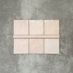 Flat lay showing Zellige Pink Mist Square 13.2 × 13.2cm Moroccan-style ceramic tiles paired with a matching Pink Mist Bullnose 3 × 20cm trim tile in soft pale pink tones, arranged on a textured grey background to highlight the glossy glaze, subtle tonal variation and handcrafted zellige-effect finish.