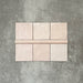 Flat lay showing Zellige Pink Mist Square 13.2 × 13.2cm Moroccan-style ceramic tiles paired with a matching Pink Mist Bullnose 3 × 20cm trim tile in soft pale pink tones, arranged on a textured grey background to highlight the glossy glaze, subtle tonal variation and handcrafted zellige-effect finish.