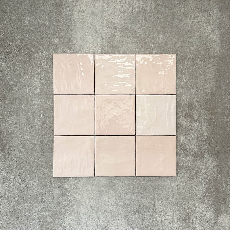 Flat lay of Zellige Pink Mist Square 13.2 × 13.2cm Moroccan-style ceramic tiles in soft pale pink and blush tones, arranged in a simple grid on a textured grey background, showcasing the glossy glaze, subtle tonal variation and handcrafted zellige-effect finish.