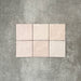 Flat lay of Zellige Pink Mist Square 13.2 × 13.2cm Moroccan-style ceramic tiles in soft pale pink and blush tones, arranged in a simple grid on a textured grey background, showcasing the glossy glaze, subtle tonal variation and handcrafted zellige-effect finish.