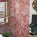 Bathroom wall featuring Zellige Rose Pink Brick 6.5 × 20cm Moroccan-style ceramic tiles, laid in a herringbone pattern with soft blush and dusty pink tones, paired with brass wall lights and taps, a green painted door and arched mirror to create a warm, characterful and handcrafted zellige-effect finish.