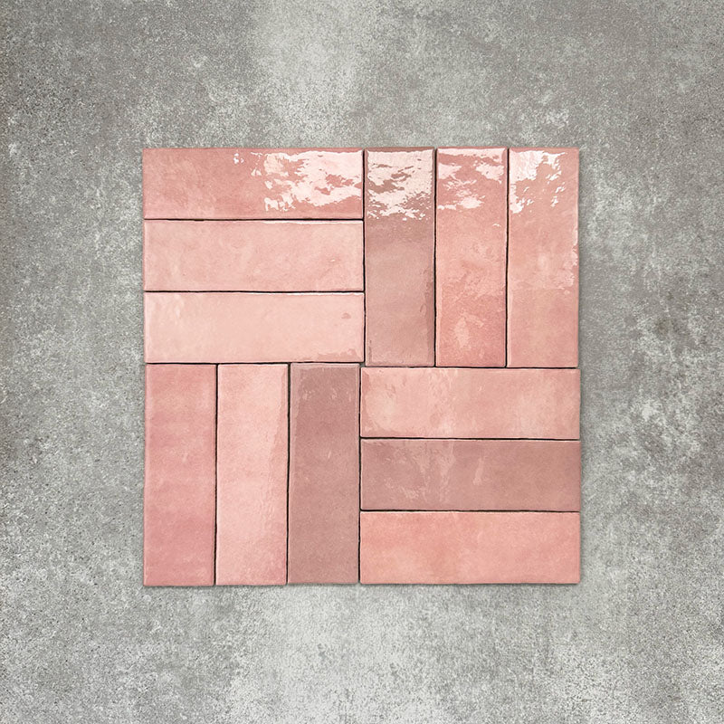 Flat lay of Zellige Rose Pink Brick 6.5 × 20cm Moroccan-style ceramic tiles, shown in soft blush, dusty rose and terracotta pink tones with gentle tonal variation and lightly uneven edges, arranged in a basketweave brick layout on a textured grey background to showcase the handcrafted zellige-effect glaze.