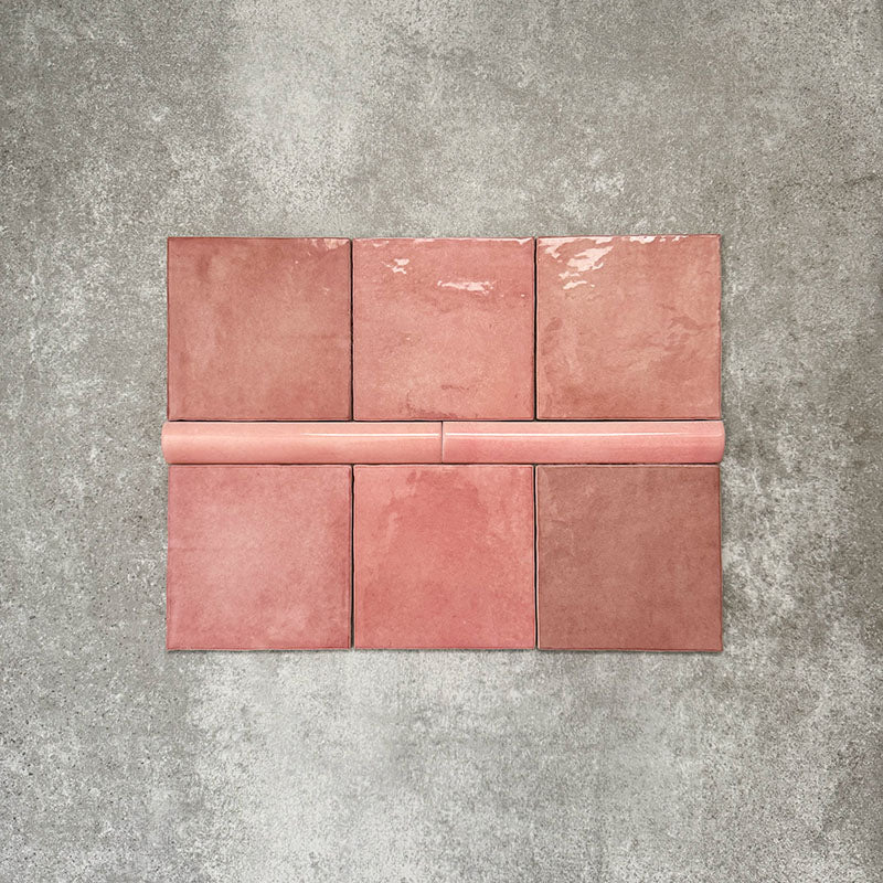 Flat lay showing Zellige Rose Pink Square 13.2 × 13.2cm Moroccan-style ceramic tiles paired with a matching Rose Pink Bullnose 3 × 20cm trim tile, displayed in soft blush and dusty pink tones with natural glaze variation and gently uneven edges, arranged on a textured grey background to demonstrate the coordinating square tile and rounded-edge bullnose detail for a handcrafted zellige-effect finish.