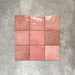 Flat lay of Zellige Rose Pink Square 13.2 × 13.2cm Moroccan-style ceramic tiles in soft blush, dusty rose and warm pink tones, showing subtle shade variation and gently uneven edges, arranged in a simple grid on a textured grey background to highlight the handcrafted zellige-effect surface.