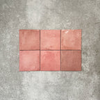 Flat lay of Zellige Rose Pink Square 13.2 × 13.2cm Moroccan-style ceramic tiles in soft blush, dusty rose and warm pink tones, showing subtle shade variation and gently uneven edges, arranged in a simple grid on a textured grey background to highlight the handcrafted zellige-effect surface.