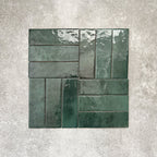 Flat lay of Zellige Royal Green Brick 6.5 × 20cm Moroccan-style ceramic tiles in rich deep green tones, arranged in a basketweave layout on a textured grey background, showcasing the glossy glaze, natural tonal variation and handcrafted zellige-effect finish.