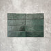 Flat lay of Zellige Royal Green Brick 6.5 × 20cm Moroccan-style ceramic tiles in rich deep green tones, arranged in a horizontal brick layout on a textured grey background, showcasing the glossy glaze, natural tonal variation and handcrafted zellige-effect finish.