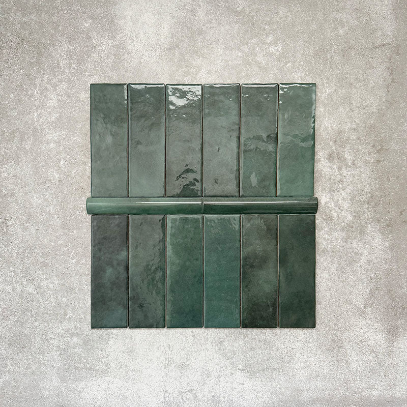 Flat lay showing Zellige Royal Green Brick 6.5 × 20cm Moroccan-style ceramic tiles paired with a matching Royal Green Bullnose 3 × 20cm trim tile in rich deep green tones, arranged on a textured grey background to highlight the glossy glaze, subtle tonal variation and handcrafted zellige-effect finish.