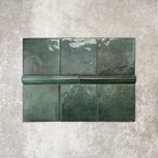 Flat lay showing Zellige Royal Green Square 13.2 × 13.2cm Moroccan-style ceramic tiles paired with a matching Royal Green Bullnose 3 × 20cm trim tile in rich deep green tones, arranged on a textured grey background to highlight the glossy glaze, subtle tonal variation and handcrafted zellige-effect finish.