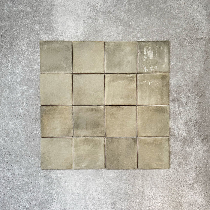 Flat lay of Zellige Weald Green 10×10cm Moroccan-style ceramic square tiles, highlighting soft green and earthy tonal variation, irregular edges and a handmade-look glazed finish on a neutral background.