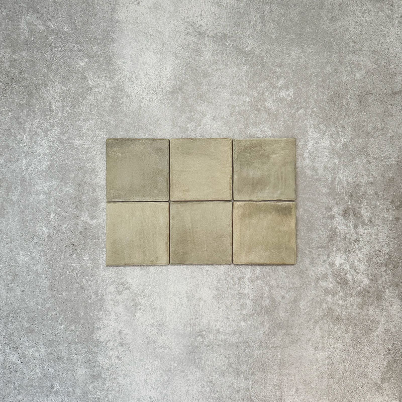 Flat lay of Zellige Weald Green 10×10cm Moroccan-style ceramic square tiles, highlighting soft green and earthy tonal variation, irregular edges and a handmade-look glazed finish on a neutral background.