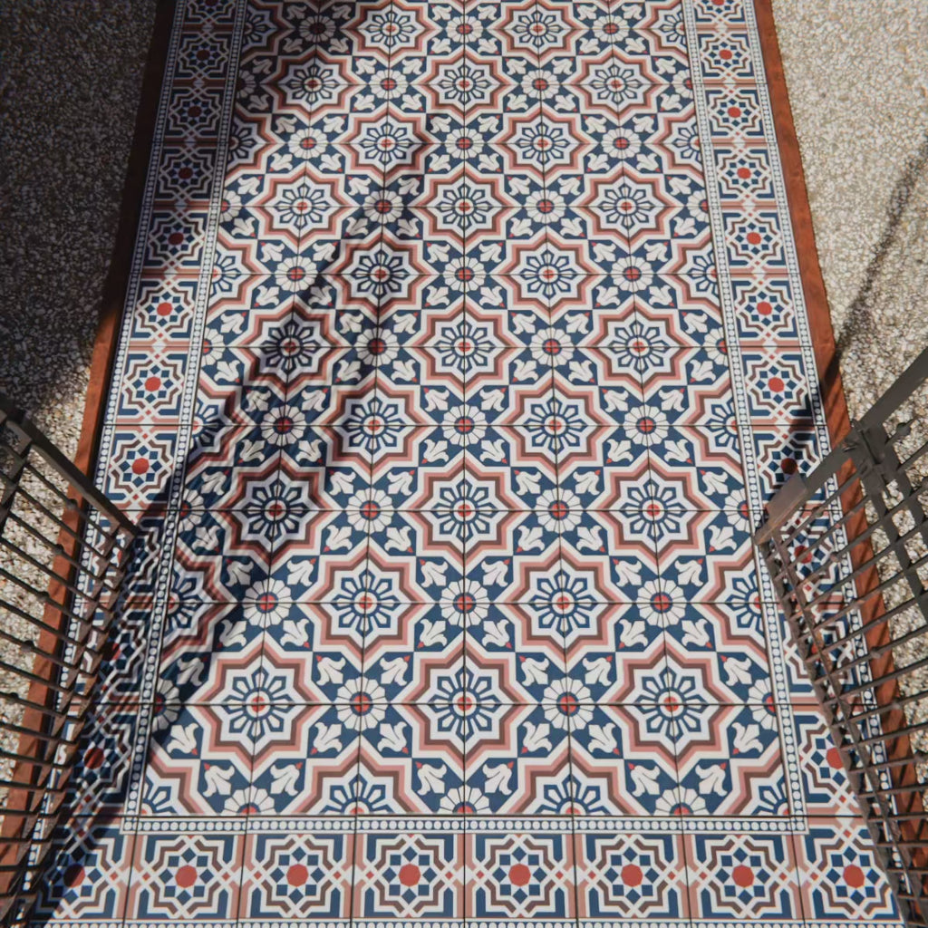 Outdoor walkway featuring Balthazar Grand 20cm x 20cm Victorian Moroccan style ceramic floor tiles, showcasing an intricate navy, terracotta and white geometric pattern, styled with black railings and natural sunlight.